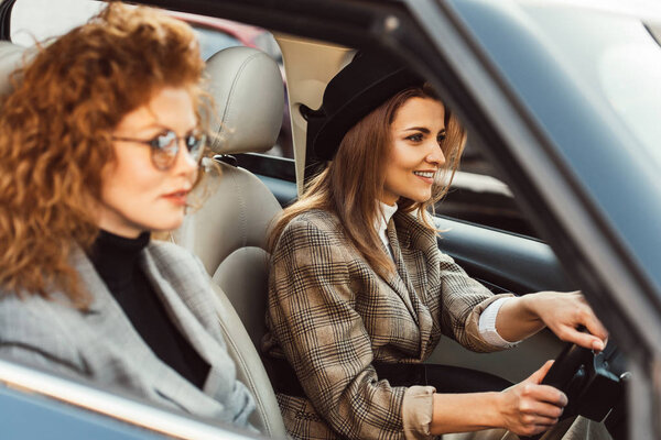 cheerful woman in black hat driving car while her ginger female friend sitting near 