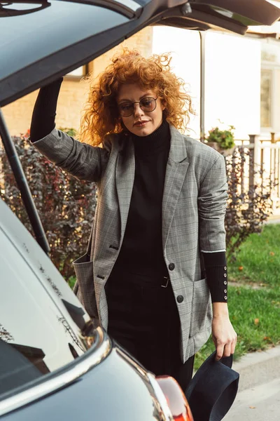 グレー ジャケットとメガネの街で車のトランクを閉じるでスタイリッシュな巻き毛の赤毛の女性 — ストック写真