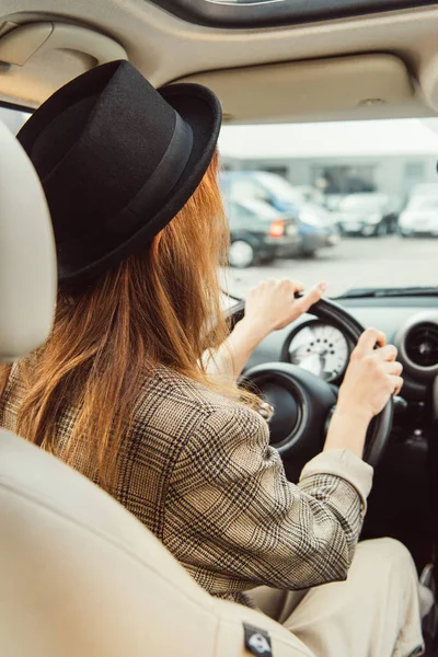 rear view of woman in black hat and checkered jacket sitting at steering wheel in car