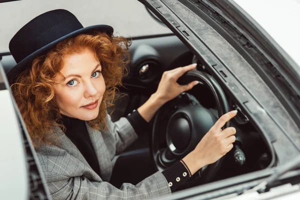 黒い帽子とステアリング ホイールの車の中で座っているグレーのジャケットでスタイリッシュな生姜女性の肖像 — ストック写真