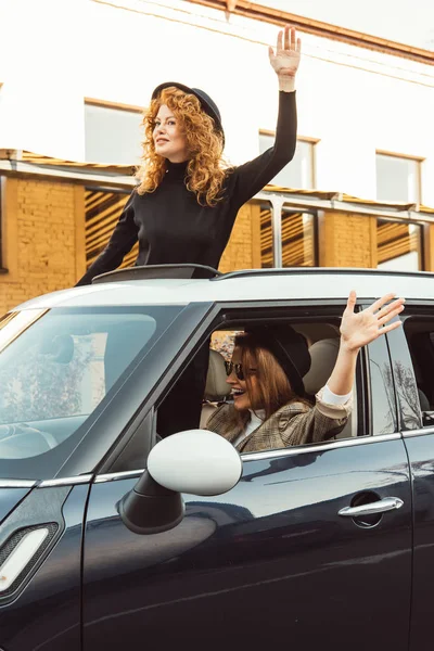 幸せな女性の友人で黒い帽子を車から身を乗り出して 都市通りで手を振って  — 無料ストックフォト