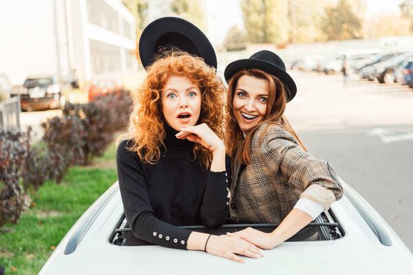 cheerful woman taking off black hat from her shocked female friend while they leaning out from car hatchway at city street