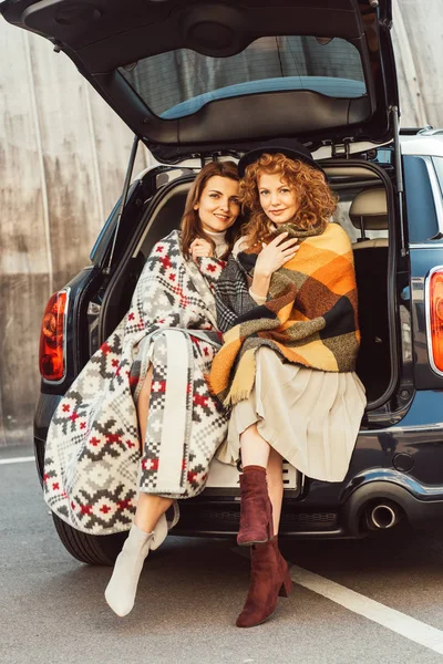 Stylish Female Friends Wrapped Blankets Looking Camera Sitting Car Trunk — Stock Photo, Image