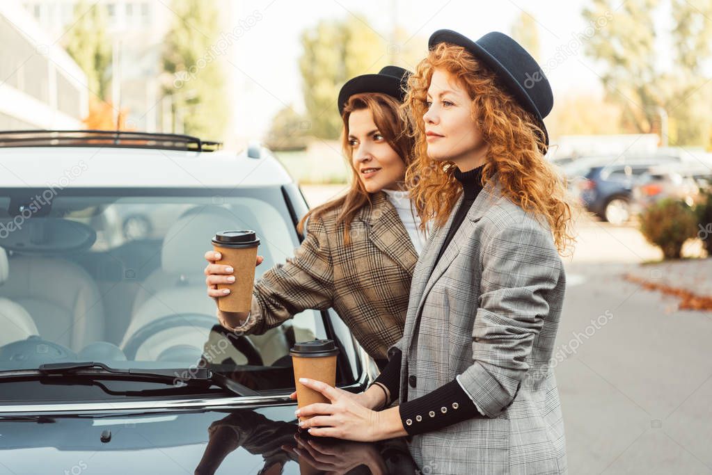 side view of stylish women in black hats and jackets standing with paper coffee cups near car at street