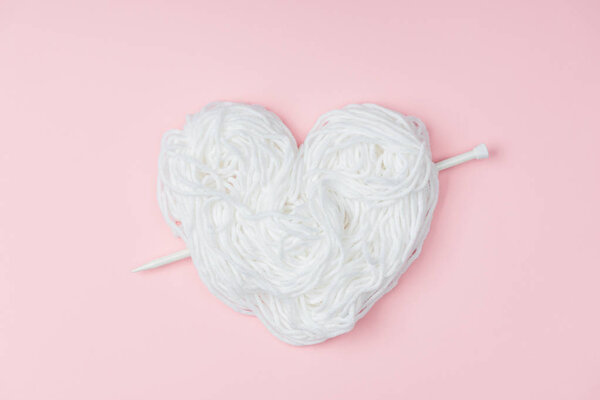 top view of yarn arranged in heart symbol and knitting needle on pink backdrop