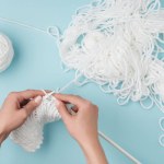 Partial view of woman with white yarn and knitting needles knitting on blue backdrop