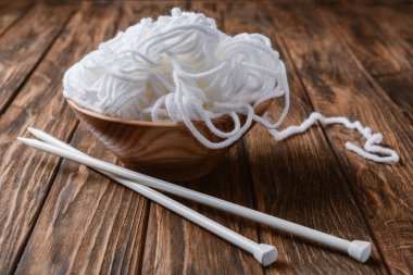 close up view of white yarn in bowl and knitting needles on wooden tabletop clipart