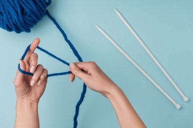 partial view of woman with blue yarn and white knitting needles knitting on blue backdrop clipart