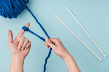 partial view of woman with blue yarn and white knitting needles knitting on blue backdrop clipart