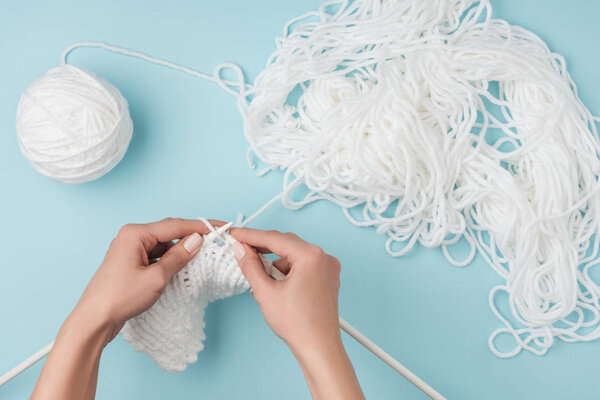 partial view of woman with white yarn and knitting needles knitting on blue backdrop