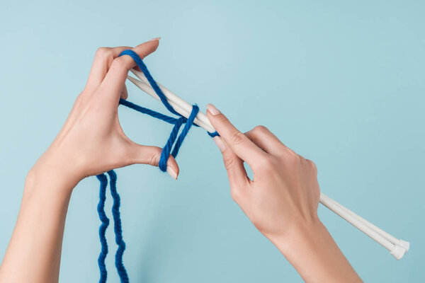 partial view of woman with blue yarn and white knitting needles knitting on blue backdrop