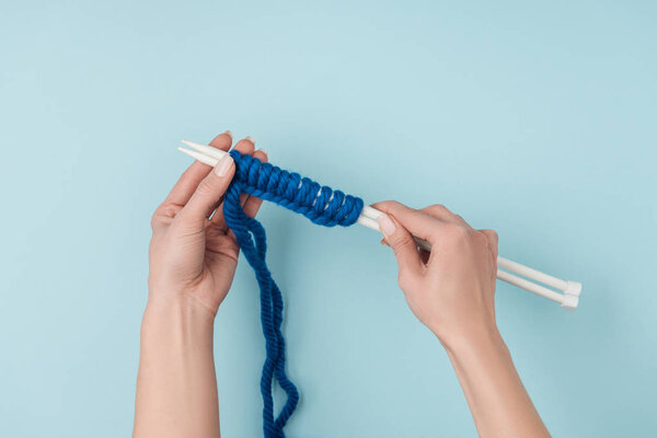 partial view of woman with blue yarn and white knitting needles knitting on blue backdrop