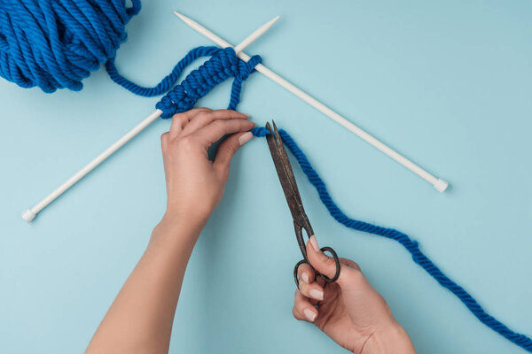 partial view of woman cutting thread with scissros on blue backdrop with yarn and knitting needles