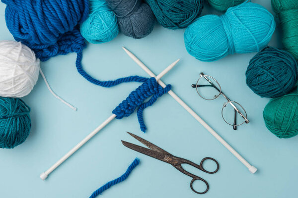 close up view of yarn, knitting needles, eyeglasses and scissors on blue backdrop
