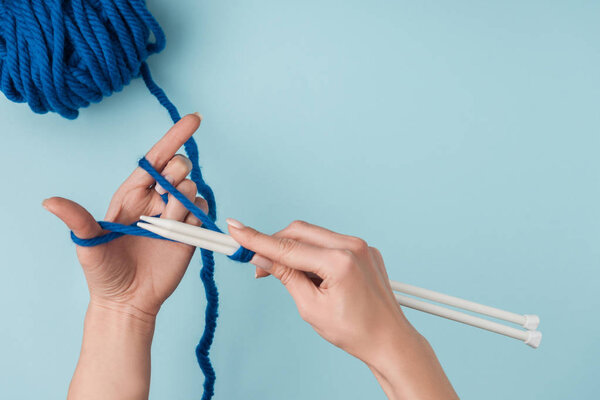partial view of woman with blue yarn and white knitting needles knitting on blue backdrop