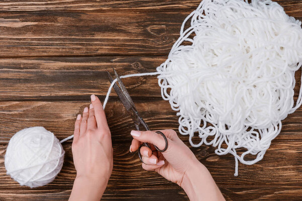 partial view of woman cutting white knitting thread with scissors on wooden surface