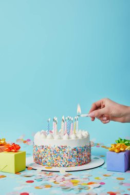 Partial view of woman lighting birthday cake on blue background with gifts and confetti clipart