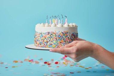 Partial view of woman holding birthday cake with candles on blue background with confetti clipart