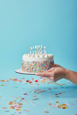 Cropped view of woman holding delicious birthday cake with candles on blue background with confetti clipart