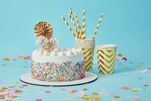 Delicious cake, plactic cups and drinking straws on blue background with confetti
