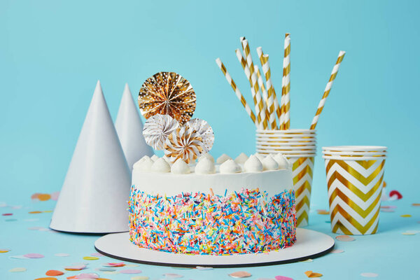 Delicious cake, plactic cups, party hats and drinking straws on blue background with confetti