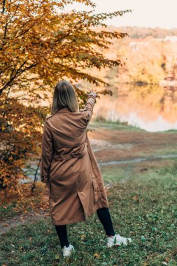 Back view of woman standing in coat and pointing at lake in autumn forest  clipart