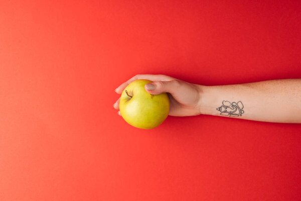 top view of tattooed female hand holding large golden delicious apple on red background