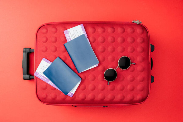 верхний вид сумки, паспорта, авиабилеты и солнцезащитные очки на красном фоне
 