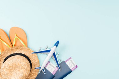 Pasaportlar, uçak bileti, hasır şapka, flip flop ve oyuncak uçak mavi zemin üzerine üstten görünüm 