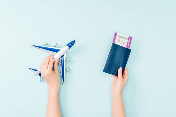 cropped view of woman holding passport, air ticket and toy plane on blue background 