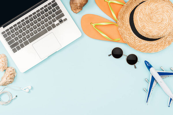 top view of toy plane, laptop with blank screen, headphones, sunglasses, seashells, flip flops, plane model and straw hat on blue background