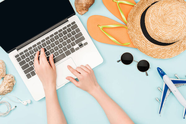 cropped view of woman using laptop near earphones, sunglasses, seashells, flip flops, toy plane and straw hat on blue background