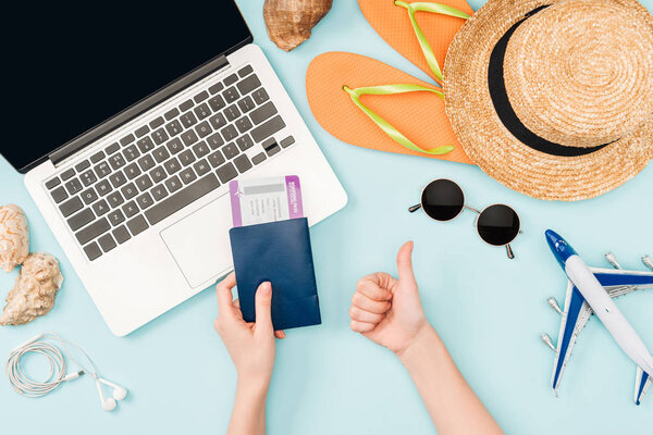 cropped view of woman holding passports and air tickets while doing thumb up gesture near laptop, earphones, sunglasses, seashells, flip flops, toy plane and straw hat on blue background