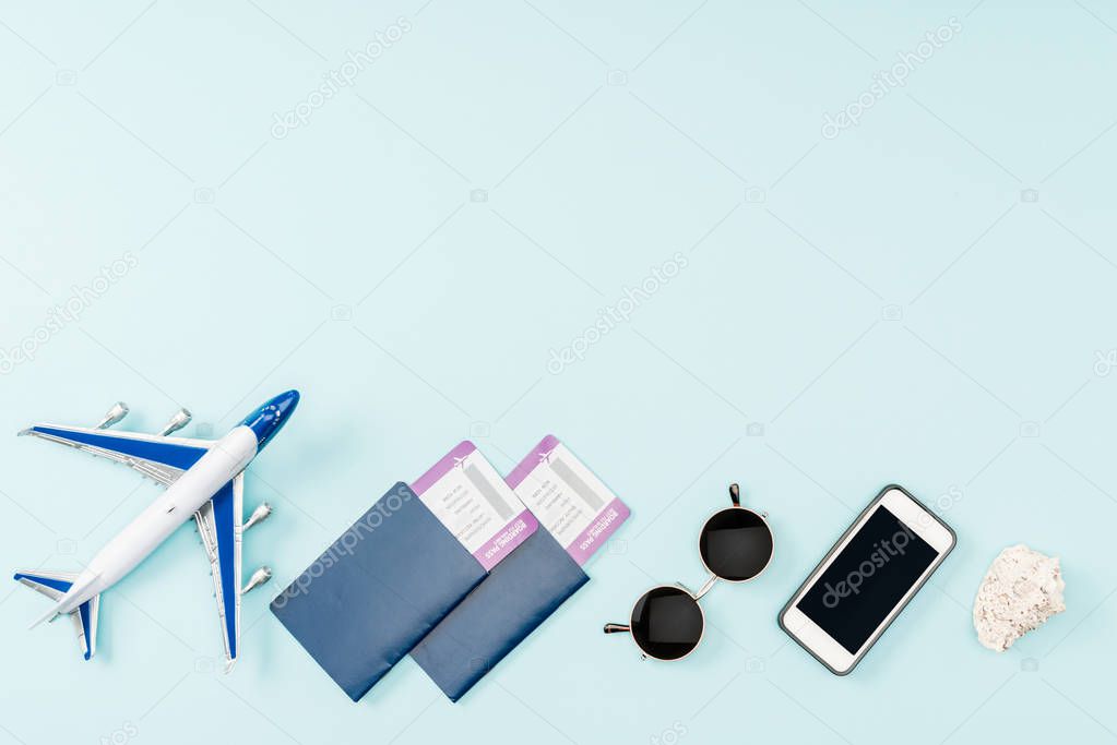 top view of passports, air tickets, toy plane smartphone with blank screen, seashells and sunglasses on blue background 
