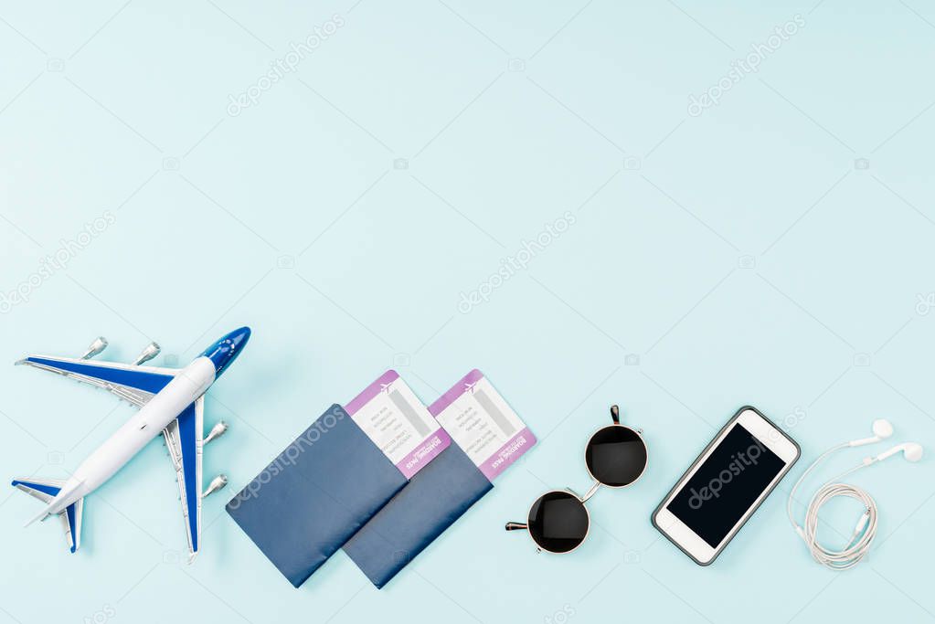 top view of passports, air tickets, toy plane, smartphone with blank screen, earphones and sunglasses on blue background 