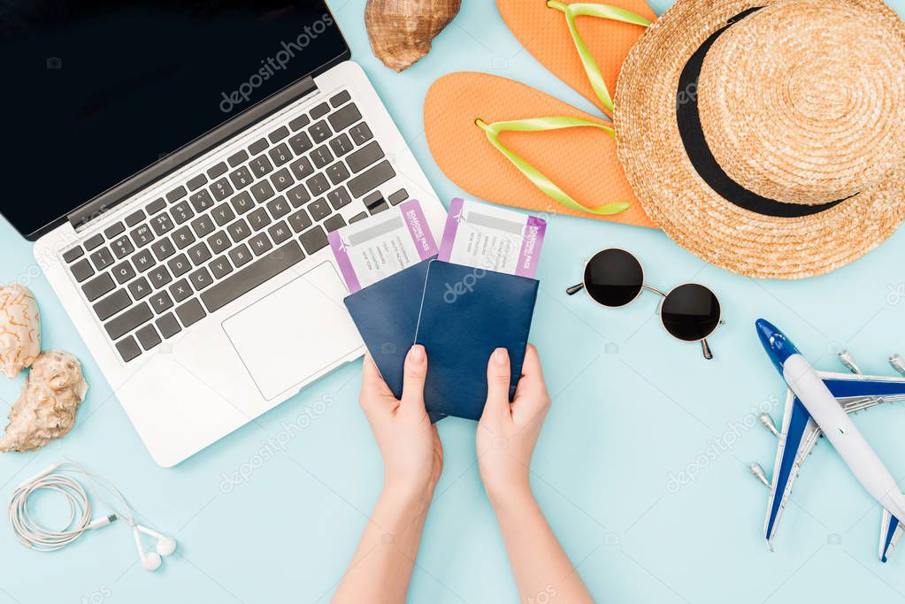cropped view of woman holding passports and air tickets near laptop, earphones, sunglasses, seashells, flip flops and straw hat on blue background