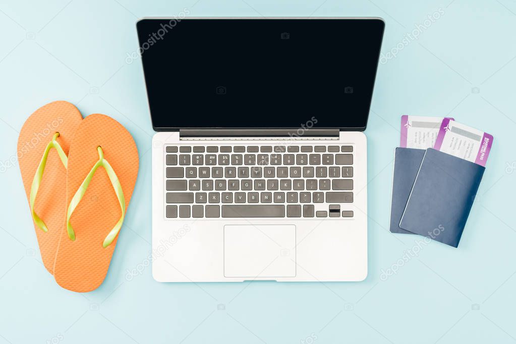 top view of laptop with blank screen, flip flops and passports with air tickets on blue background