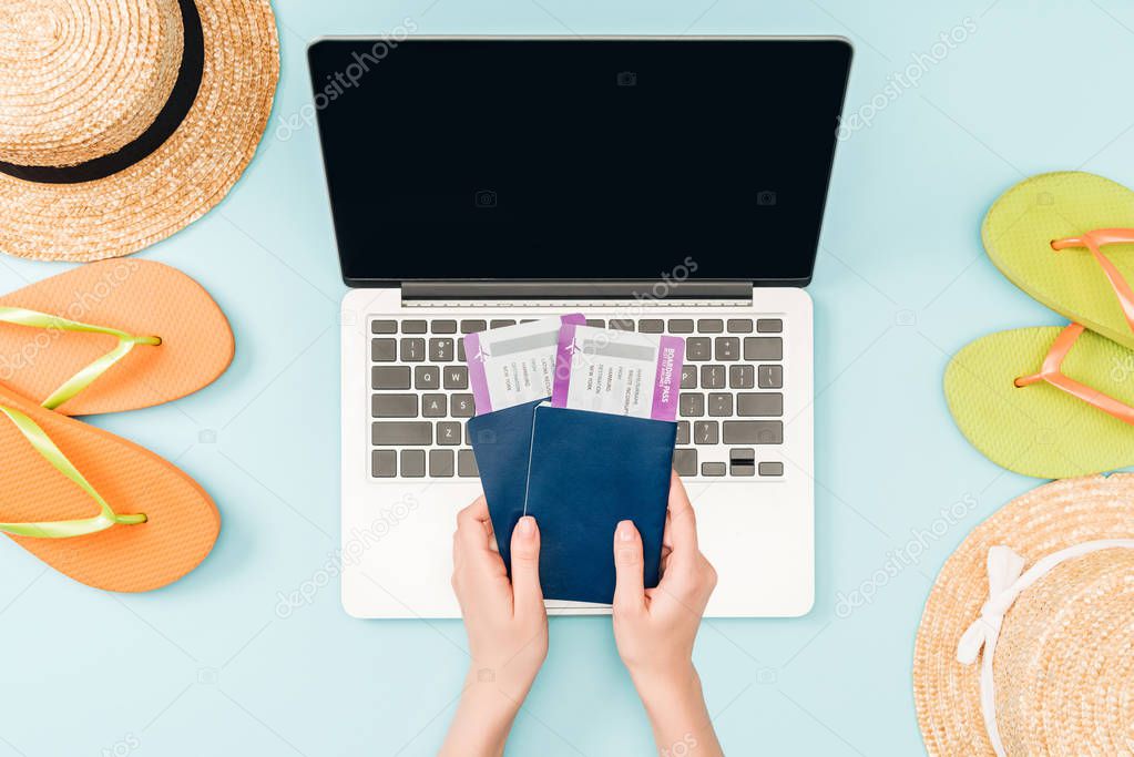 cropped view of woman holding passports with air tickets near laptop with blank screen, straw hats and flip flops on blue background