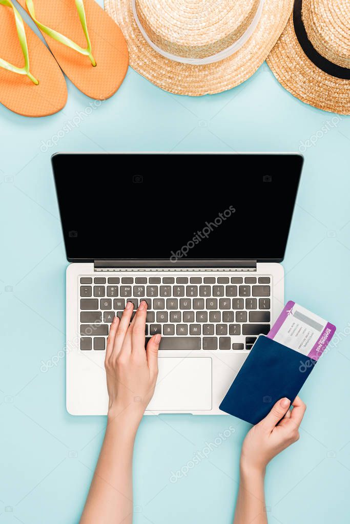cropped view of woman using laptop with blank screen and holding passport with air ticket near straw hats and flip flops on blue background