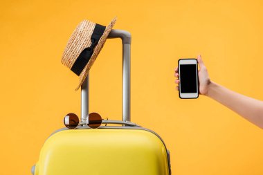 Kırpılan görünümünü smartphone ile seyahat çantası, hasır şapka ve güneş gözlüğü izole sarı yakınındaki boş ekran tutan kadın