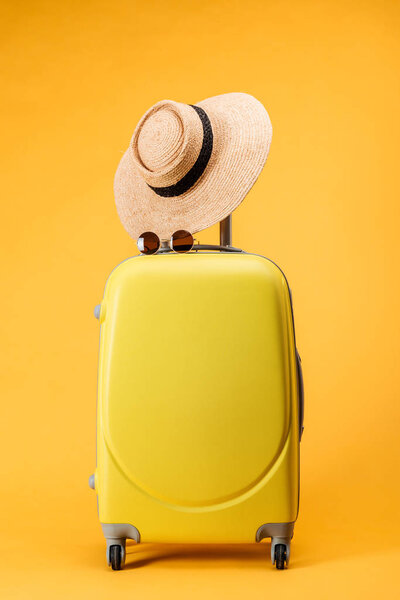 travel bag with wheels, straw hat and sunglasses on yellow background