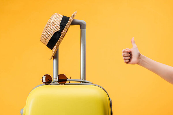cropped view of woman doing thumb up gesture near travel bag, straw hat and sunglasses isolated on yellow