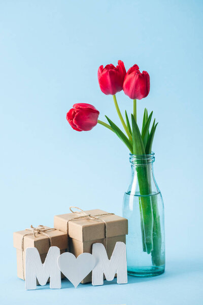 gift boxes, bouquet of red tulips and paper word mom with heart shaped litter "o" on blue background