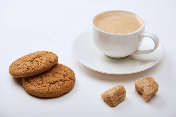 tasty coffee with foam in cup on saucer near brown sugar and cookies on white background