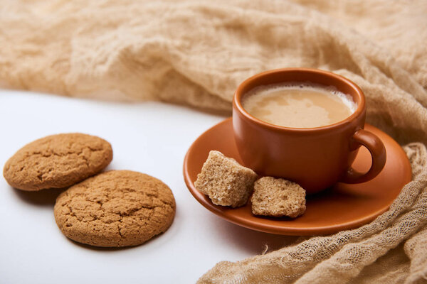 delicious coffee with foam in cup on saucer with brown sugar near cloth and cookies on white background 