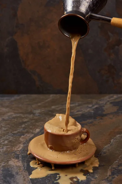 stock image pouring hot coffee from cezve into cup with puddle near saucer on marble surface