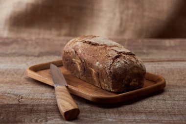 fresh brown bread loaf on chopping board near knife on wooden table clipart