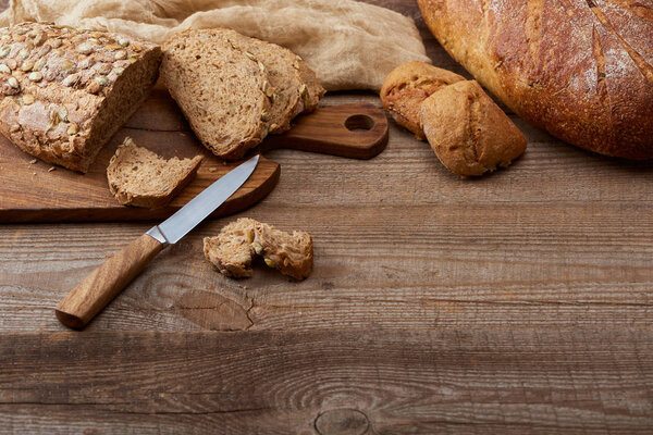 fresh cut and whole bread loaves and buns, knife and chopping board near cloth on wooden table