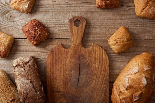 木製の素朴なテーブルの空のまな板の近くにパンとパンの新鮮な自家製パンのトップビュー — ストック写真