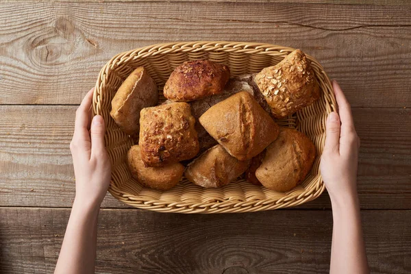 木製のテーブルの上のウィッカーバスケットに新鮮な自家製のパンを保持している女性のトップビュー — ストック写真